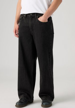 Person iført løstsiddende sorte jeans, sorte sko og en hvid skjorte, hånd i højre lomme, stående mod en ensfarvet lys baggrund.
