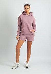 Világos rózsaszín pulóver kapucnis felső nagy elülső zsebbel, hozzáillő rövidnadrággal párosítva. Az anyag puha tapintású; hosszú ujjú és kapucnis kialakítású.