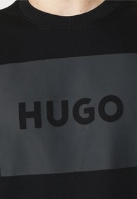 Sudadera negra con cuello redondo acanalado, que presenta un gran panel rectangular gris oscuro y letras negras en negrita que dicen "HUGO". Material de mezcla de poliéster.