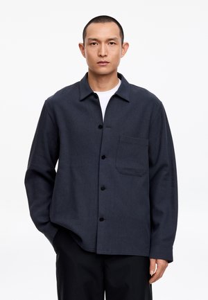 Homme aux cheveux courts portant une chemise sombre boutonnée sur un t-shirt blanc et un pantalon noir, debout devant un fond blanc.