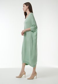Largo vestido holgado de suave tejido verde menta, con escote en V, mangas de tres cuartos y un dobladillo ligeramente curvado. Combinado con tacones altos en tono nude.