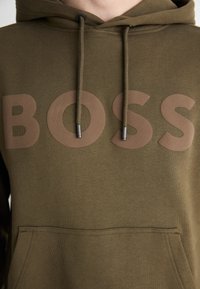 Olivgrüner Hoodie mit einem großen braunen "BOSS"-Logo auf der Vorderseite, mit Kapuze und Kängurutasche. Weicher Stoff.