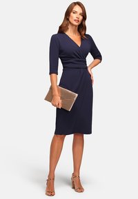 Femme en robe portefeuille bleu marine tenant une pochette métallique, portant des sandales à talons et des boucles d'oreilles pendantes, se tenant avec une main sur la hanche.