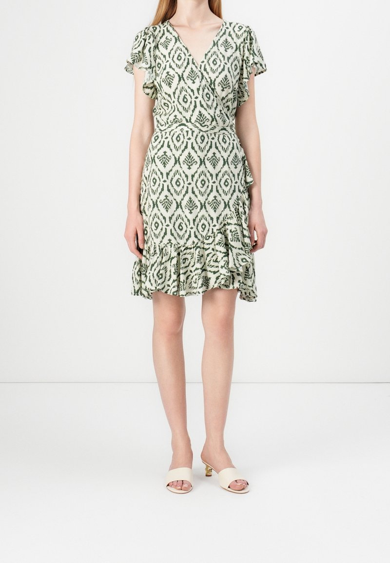Femme portant une robe portefeuille à manches courtes, à motif vert et blanc, avec un ourlet froncé et des sandales beiges à talons ouvertes.