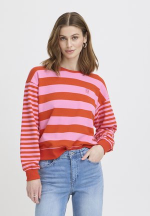 Noor naine õlgadeni juustega, kannab roosa ja punase triibulise kampsunit ning helerohelisi teksapükse, poseerides ühe käega taskus.