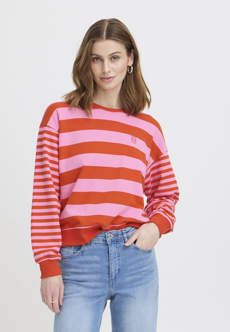 Mladá žena s vlasy po ramena, oblečená v růžovém a červeném pruhovaném svetru a světlých modrých džínách, pózující s jednou rukou v kapse.