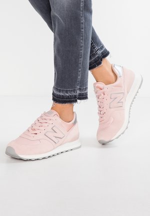 Osoba nosząca różowe sneakersy New Balance z szarymi akcentami oraz ciemne przycięte dżinsy stojąca na białej powierzchni.