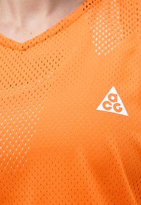 Κοντινό πλάνο πορτοκαλί αθλητικής μπλούζας με τρύπες και λευκό λογότυπο ACG στην περιοχή γιακά στο στήθος.