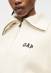 Κοντινό πλάνο ενός ατόμου που φοράει λευκό πλεκτό πουλόβερ με λαιμόκοψη μισού φερμουάρ και κεντημένο λογότυπο "GAP" στο στήθος.