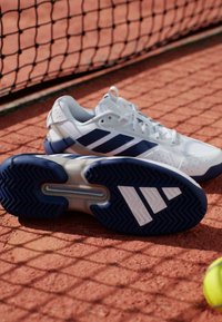 Chaussures de tennis blanches avec des accents bleu marine, dotées d'une semelle texturée, d'un design à trois bandes et d'un col rembourré. Visible sur un court en terre battue.