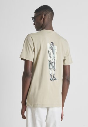 Uomo che indossa una maglietta beige con un disegno in bianco e nero di una persona in piedi sulla schiena, abbinata a pantaloni bianchi e occhiali.