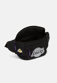 Juodas fanny packas, pagamintas iš patvarios medžiagos, su Los Andželo Lakers logotipu ir baltu grafikiniu tekstu, papuoštas violetiniais užtrauktukais ir akcentais.