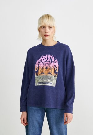 Sweatshirt bleu marine en tissu texturé avec une impression graphique colorée de vagues et de palmiers, accompagnée du texte "SURFING SINCE 1990."