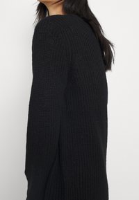 Svart tröja med ett strukturerat stickmönster, långa ärmar och en avslappnad passform, som visar en sidovy av plaggets form.