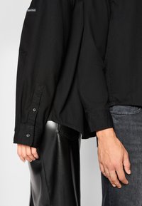 Two people stand side by side, one wearing a black shirt and leather pants, the other wearing a black shirt and dark gray jeans.