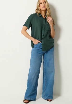 Femme portant une chemise à manches courtes boutonnée vert foncé, un jean large bleu clair et des sandales foncées, debout contre un mur blanc uni.