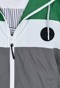 Kurtka z blokowym kolorowaniem w kolorach zielonym, białym i szarym, z zapięciem na zamek, sznurkiem regulacyjnym oraz okrągłą naszywką z logo na klatce piersiowej.