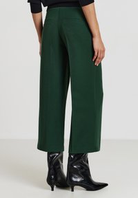 Pantaloni a gamba larga in tessuto verde scuro, con una texture liscia e tasche posteriori. Abbinati a stivaletti neri lucidi.