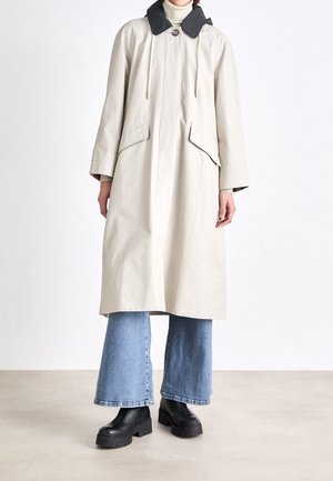Le mannequin porte un long manteau crème à col foncé, un jean bleu à jambes larges et des bottes noires à plateforme.