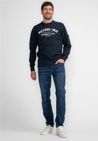 Donkerblauwe sweatshirt met "PETROL IND ALASKA U.S.A" in het wit gedrukt. Gecombineerd met blauwe jeans en witte sneakers. Zachte stof, casual pasvorm.