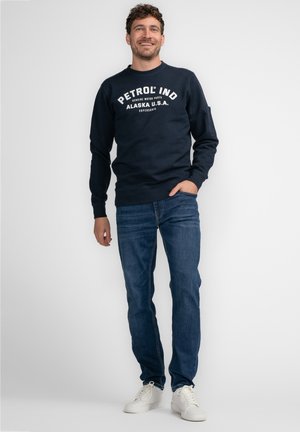 Sudadera azul marino con "PETROL IND ALASKA U.S.A" impreso en blanco. Combinada con jeans azules y zapatillas blancas. Tela suave, corte casual.
