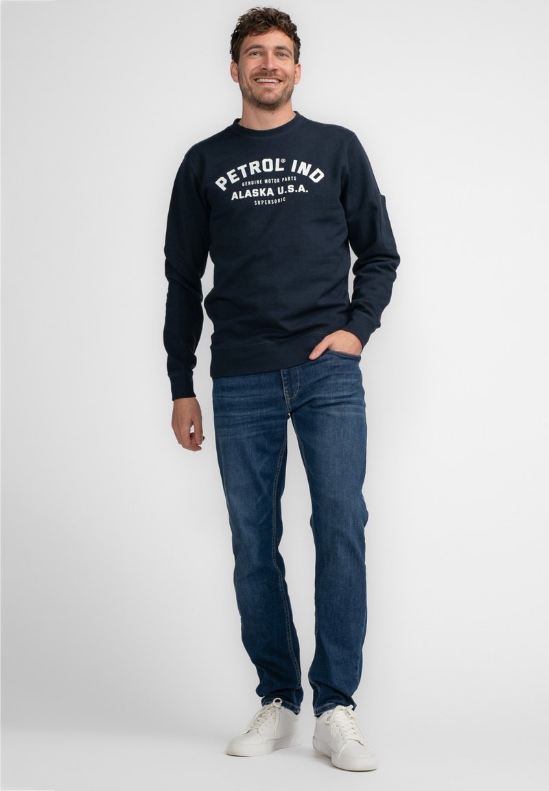 Donkerblauwe sweatshirt met "PETROL IND ALASKA U.S.A" in het wit gedrukt. Gecombineerd met blauwe jeans en witte sneakers. Zachte stof, casual pasvorm.