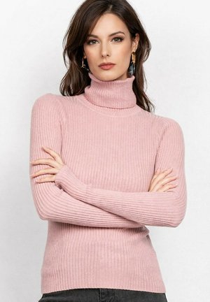 Femme aux longs cheveux bruns portant des boucles d'oreilles dorées et noires, vêtue d'un pull à col roulé côtelé rose, les bras croisés, fond neutre.