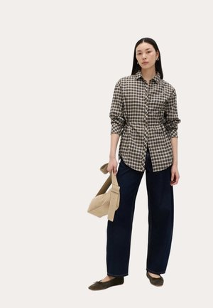 Frau trägt ein langärmeliges beige-schwarz kariertes Hemd, dunkelweite Hose, braune Ballerinas, hält eine beigefarbene Handtasche mit breitem Träger in der rechten Hand.