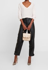 Beige texturerad handväska med lock och guldfärgad hårdvara. Modell i ljus V-ringad tröja, svarta läderbyxor och högklackade skor med ormskinnsmönster.