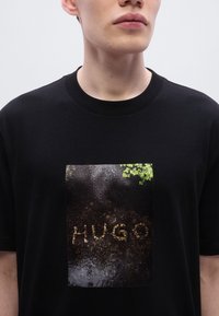 Μαύρο βαμβακερό T-shirt με γυαλιστερό σχέδιο εδάφους όπου η λέξη «HUGO» σχηματίζεται από πέτρες, με λεπτομέρειες πράσινων φυτών.
