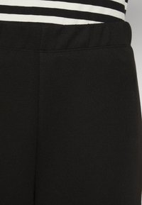 Leggings noirs en mélange de coton avec une taille élastique, présentant une texture lisse et des détails de couture minimaux. Un tissu rayé est visible au-dessus.