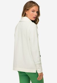 Cardigan bianco a maniche lunghe con colletto, caratterizzato da una vestibilità rilassata e dettagli con zip laterale, abbinato a pantaloni verde aderenti.