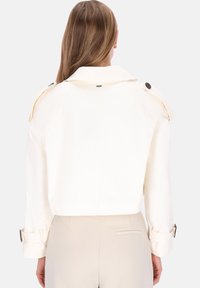 Cremfarbener, gekürzter Trenchcoat mit Schulterklappen, großen Knöpfen und aufgerollten Manschetten. Glatte Textur, minimalistisches Design.