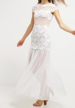 Kobieta w białej koronkowej sukience z kwiatowymi wzorami i przezroczystą tkaniną w groszki, w połączeniu z beżowymi sandałami na blokowym obcasie z paskami.