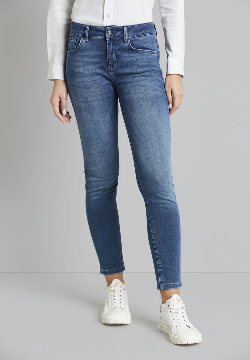 Skinny jeans in medium blue denim with five pockets, stitched in contrasting white. Worn with white sneakers featuring textured soles.