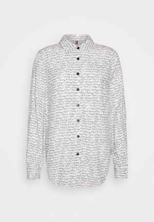 Chemise blanche à manches longues avec motif texte en cursive noire ""tommy hilfiger"" et boutons noirs sur le devant, présentée sur fond uni.