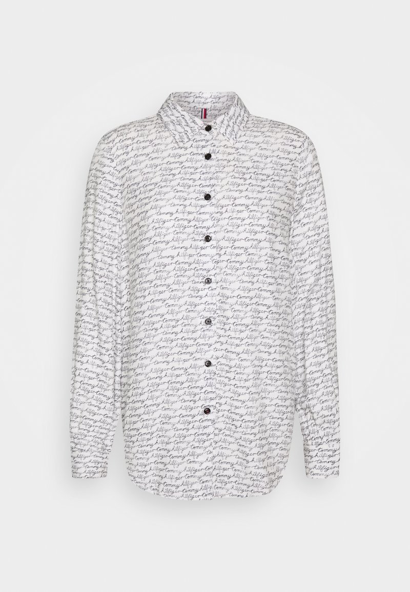 Chemise blanche à manches longues avec motif texte en cursive noire ""tommy hilfiger"" et boutons noirs sur le devant, présentée sur fond uni.