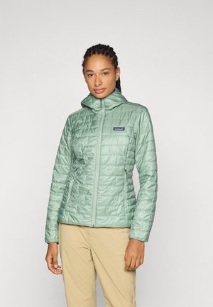 Žena s pletenými vlasmi, oblečená v svetlozelenom prešívanom kabáte s kapucňou a béžovými nohavicami, stojaca pred jednoduchým svetlým pozadím.