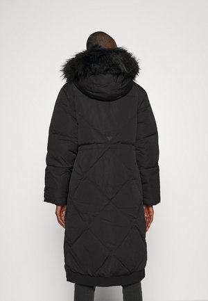 Personne portant un long manteau d'hiver matelassé noir avec une capuche bordée de fourrure noire, vue de dos sur un fond uni.