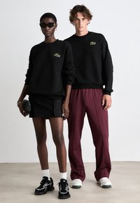 Crni duksevi s logotipom Lacoste; model nosi crnu plisiranu suknju; bordo hlače; tenisice s bijelim i zelenim detaljima; naočale za sunce.