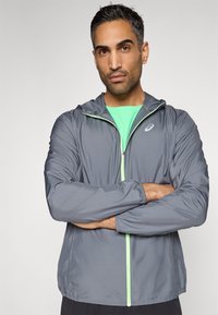 Γκρι ελαφρύ μπουφάν με κουκούλα, με φερμουάρ μπροστά και πράσινες λεπτομέρειες, φορεμένο πάνω από πράσινο πουκάμισο. Ελαστική, λεία υφή.