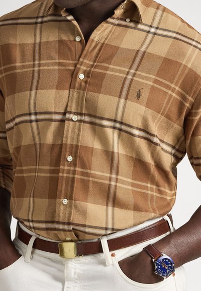 Rjava in tan karirana srajca z ovratnikom z gumbi, kombinirana z rjavim usnjenim pasom in modro ročno uro. Vidne bele hlače.