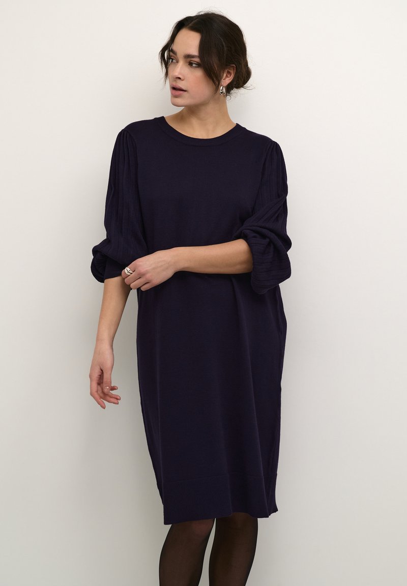 Robe tricotée bleu marine avec une coupe décontractée, mettant en avant des manches bouffantes et un ourlet côtelé. Le matériau semble doux avec une texture lisse.