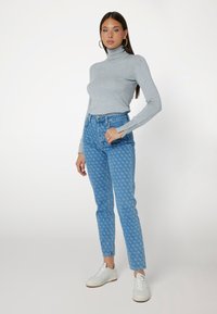 Maglione grigio a collo alto abbinato a jeans a vita alta blu con motivo e sneakers bianche, che mettono in evidenza un design aderente e una silhouette sartoriale.