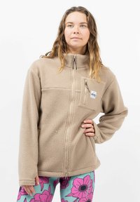 Beige Fleecejacke mit hohem Kragen und Reißverschluss vorne. Verfügt über eine Brusttasche und ein strukturiertes Material. Das Model trägt gemusterte Leggings.