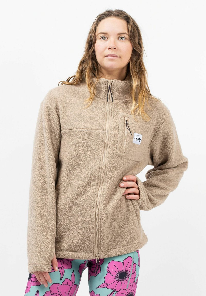 Beige Fleecejacke mit hohem Kragen und Reißverschluss vorne. Verfügt über eine Brusttasche und ein strukturiertes Material. Das Model trägt gemusterte Leggings.