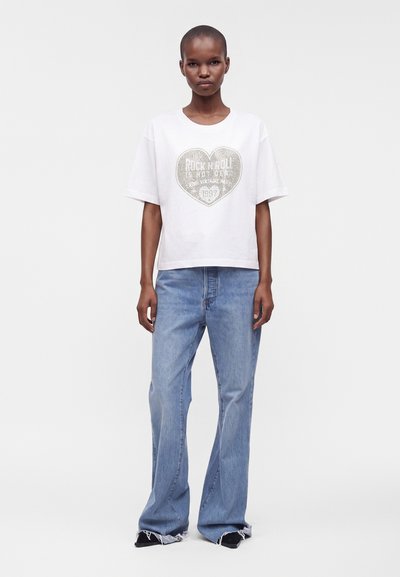 Woman wearing a white short-sleeve t-shirt with a heart and text, paired with light blue wide-leg jeans and black shoes, standing against white background.