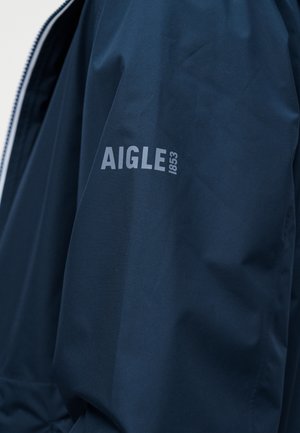 Marineblå jakkeerm med logoet "AIGLE 1853" trykt i lysegrått nær øvre armområde.