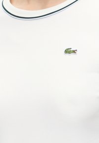 Chemise blanche avec un col rond, une bordure vert foncé et un petit logo crocodile vert brodé sur la poitrine gauche.