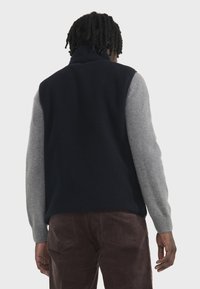 Gilet en polaire marine avec des manches tricotées grises, col haut et poignets côtelés. Présente un design ajusté et un tissu texturé.